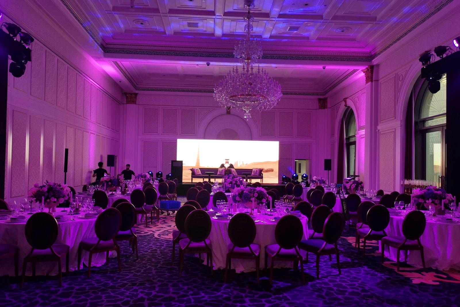 A ballroom dressed for a gala dinner, lit in magenta and blue, with round tables and an LED stage